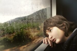 child sat on a train staring out of the window, looking sad